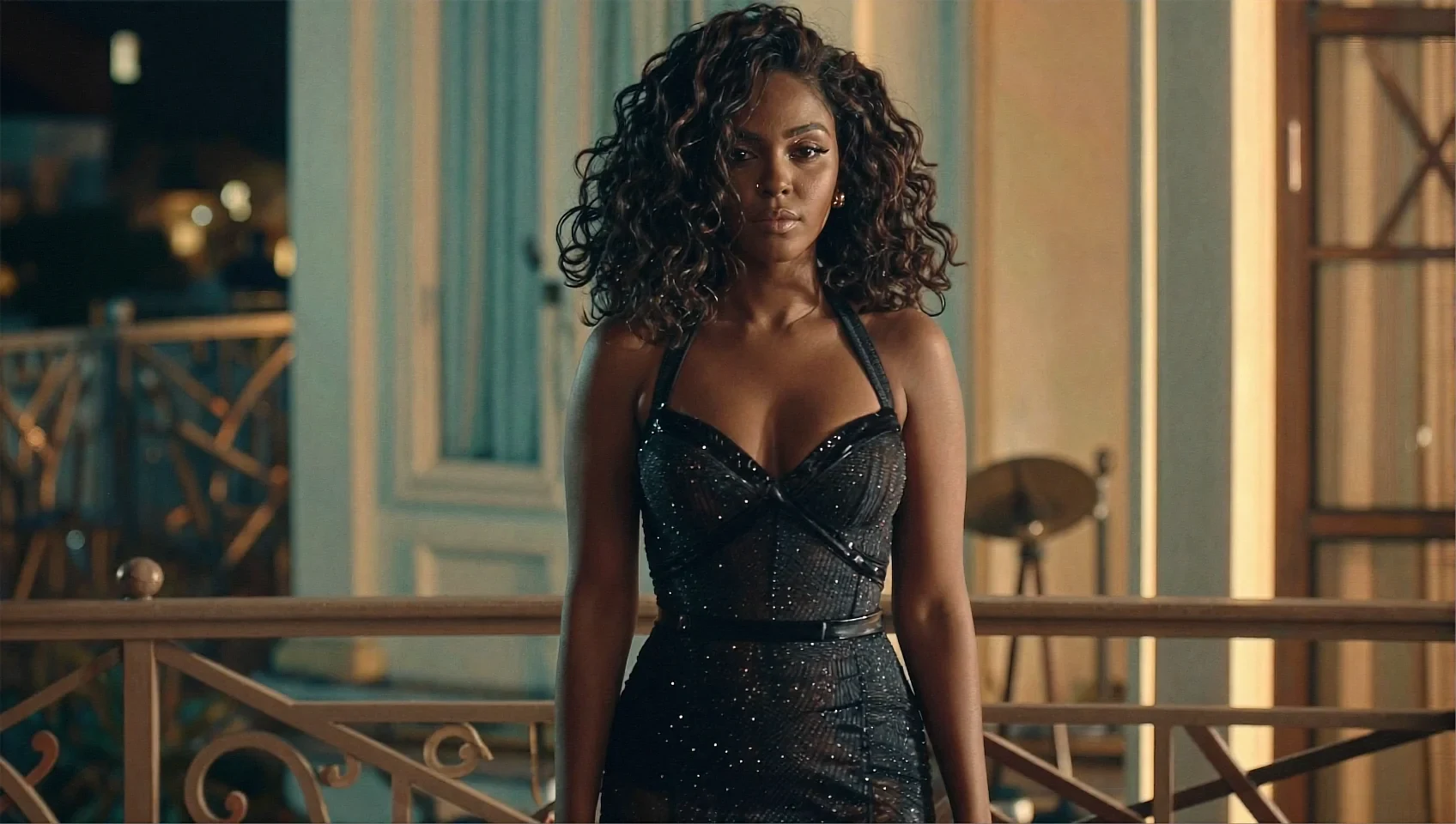 Woman with curly dark hair in sparkly black halter bustier dress standing on ornate balcony with warm vintage lighting